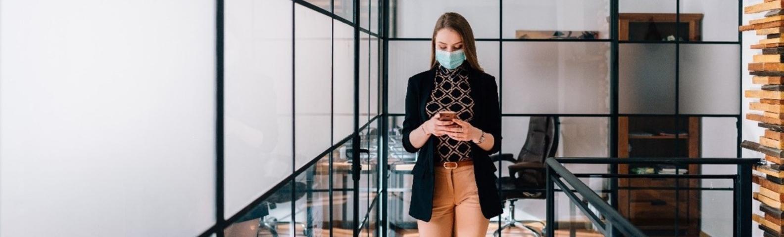 A woman texting in an office as she wears a mask